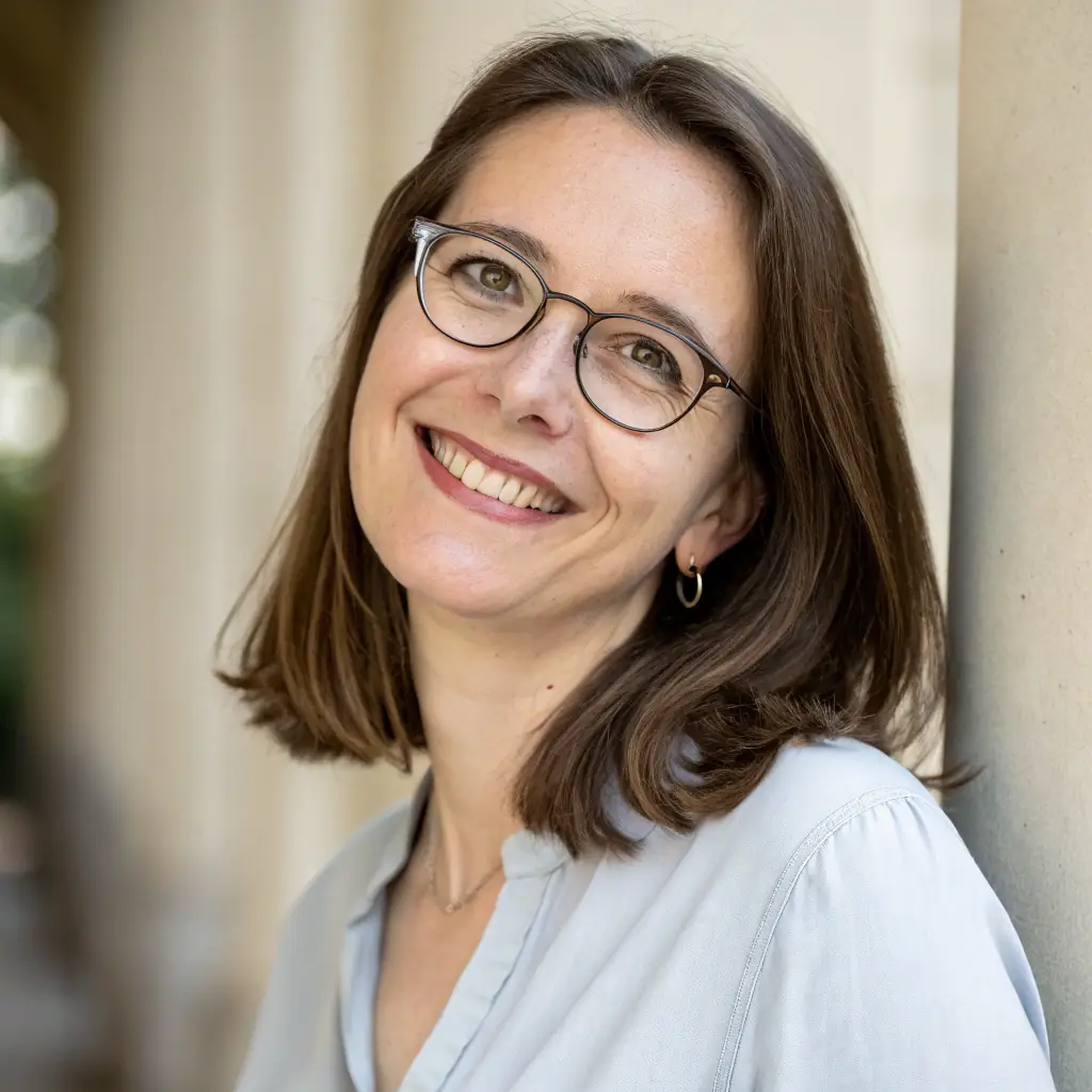 Smiling woman wearing glasses, leaning against a wall in a softly lit outdoor setting